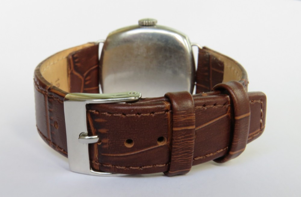 A 1930s cushion-shaped stainless steel wristwatch featuring a light dial with black Arabic numerals, a red twelve marker, a subsidiary seconds dial, and a textured brown leather strap. View V