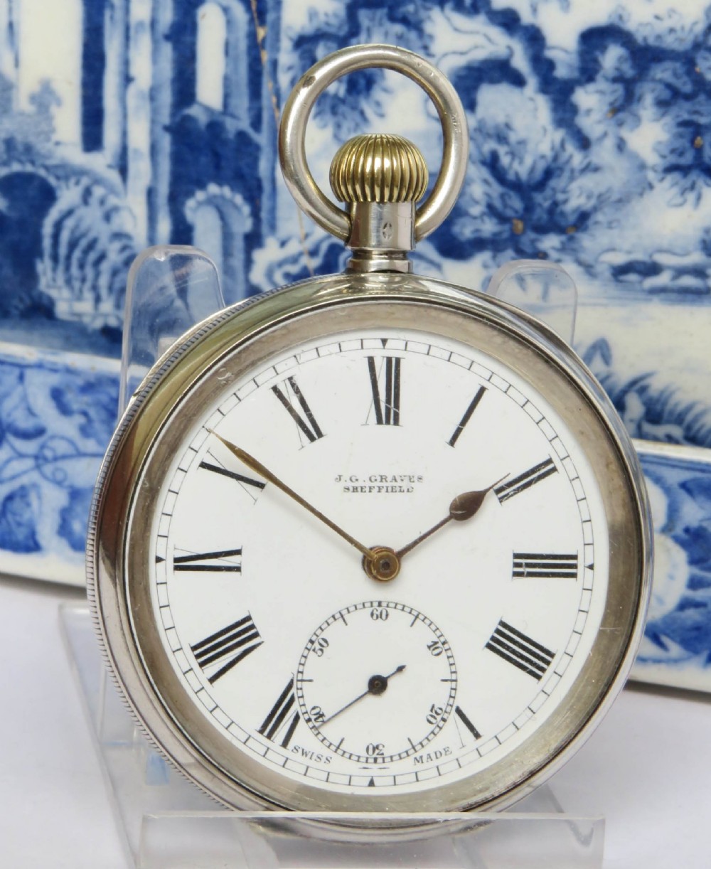 An open-face silver pocket watch featuring a white enamel dial with black Roman numerals, a sub-seconds dial at the bottom, and a brass winding crown at the top. View II