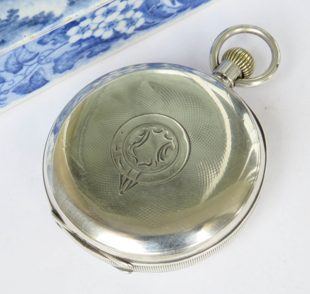 An open-face silver pocket watch featuring a white enamel dial with black Roman numerals, a sub-seconds dial at the bottom, and a brass winding crown at the top. View V