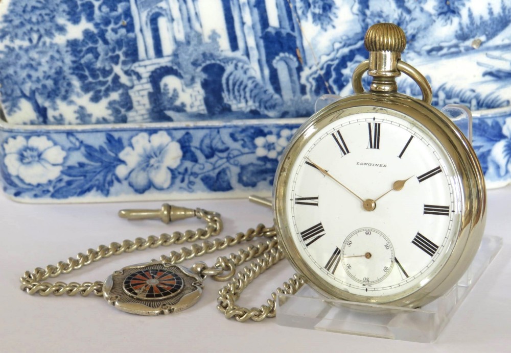 An antique open-face Longines pocket watch with a white enamel Roman numeral dial, connected to a silver-coloured curb chain with an enamelled fob medallion.
