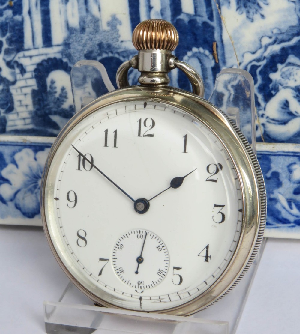 An open-face antique silver pocket watch featuring a white enamel dial with black Arabic numerals, a sub-seconds dial, and a ribbed gold-toned crown.