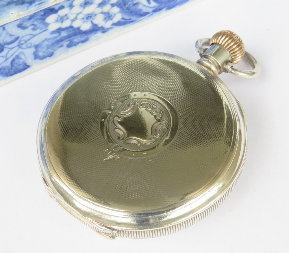 An open-face antique silver pocket watch featuring a white enamel dial with black Arabic numerals, a sub-seconds dial, and a ribbed gold-toned crown. View V