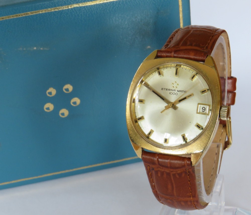 A 1969 gents Eterna-Matic 1000 wristwatch featuring a gold-toned cushion case, champagne dial with date aperture, and brown leather strap.