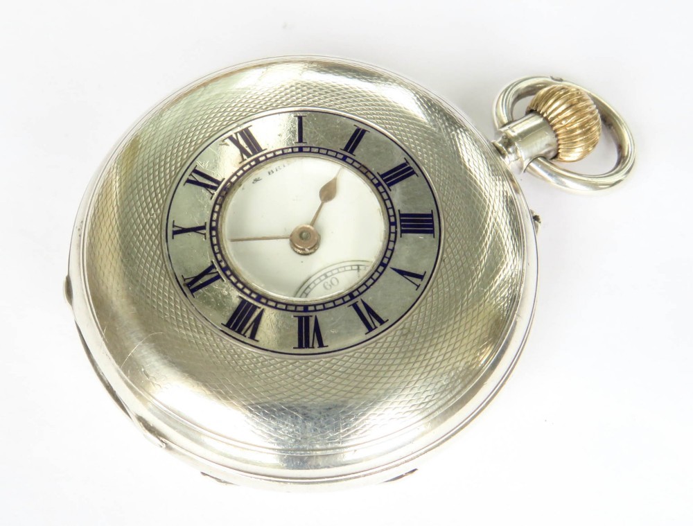 An antique sterling silver half hunter pocket watch featuring an engine-turned case with blue enamel Roman numerals around a central aperture.
