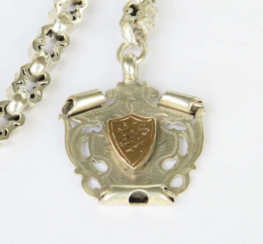 An antique sterling silver pocket watch chain featuring decorative carved links, a T-bar attachment, a swivel clasp, and a shield-shaped fob with gold detailing. View II