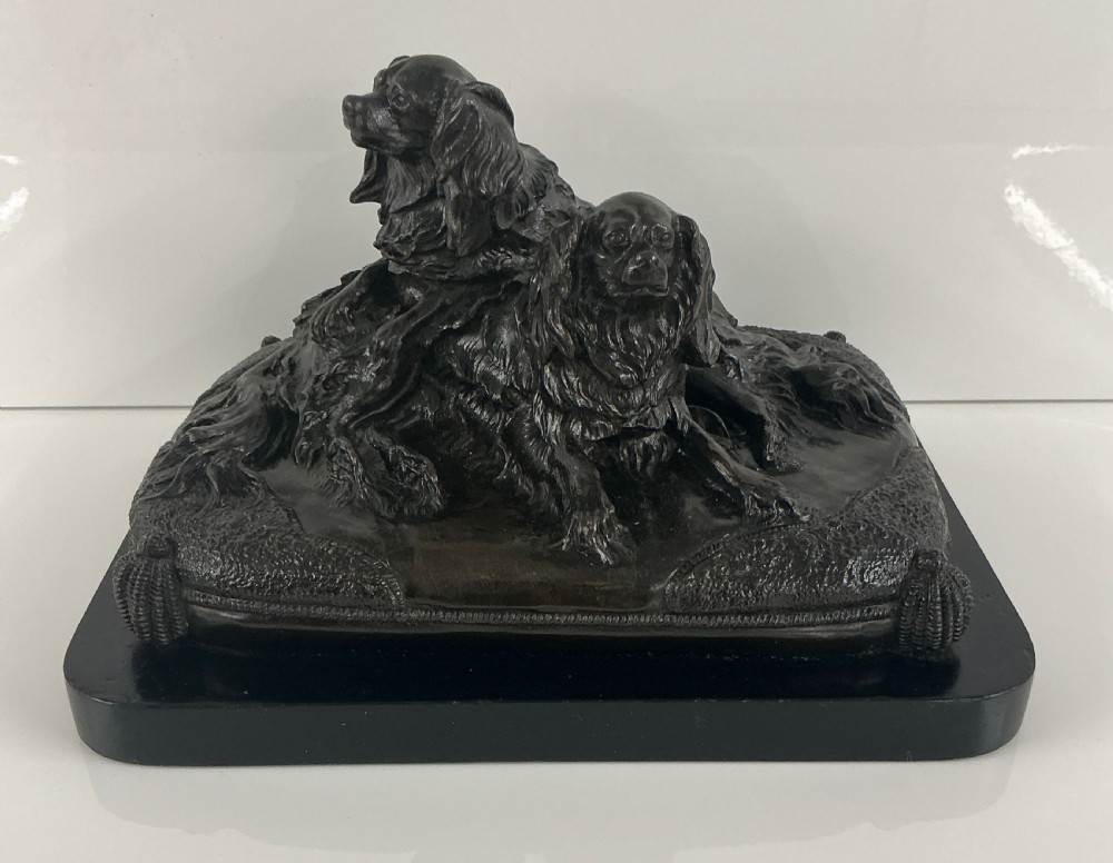 A dark patinated French bronze sculpture depicting two King Charles Spaniels resting on a tasseled cushion mounted on a black base.
