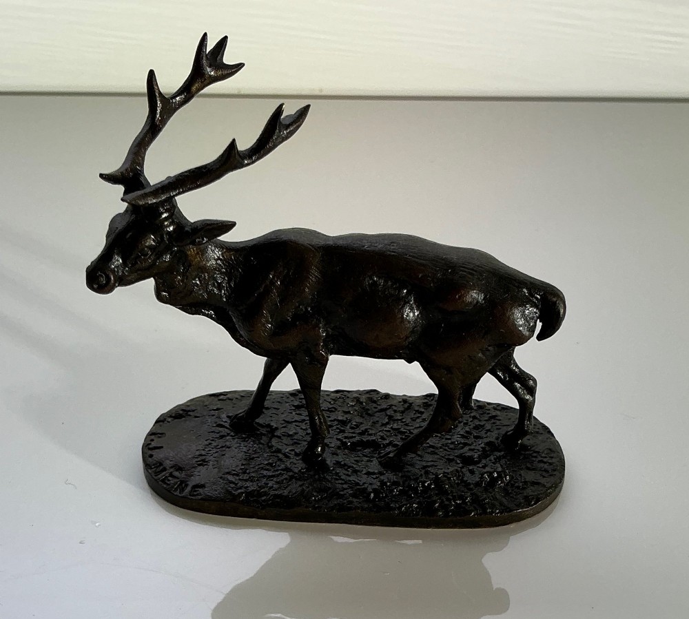A dark patinated bronze sculpture of a standing stag with multi-pointed antlers, set on an oval textured base bearing a signature.