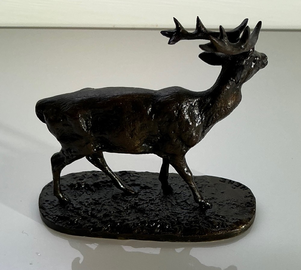 A dark patinated bronze sculpture of a standing stag with multi-pointed antlers, set on an oval textured base bearing a signature. View II