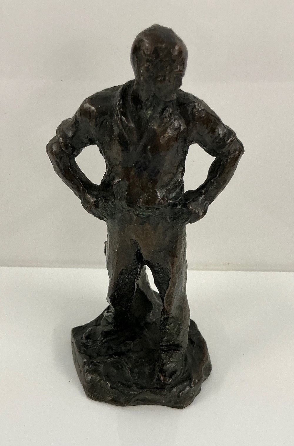 A dark patinated bronze sculpture depicting a standing male worker with his hands resting on his hips.