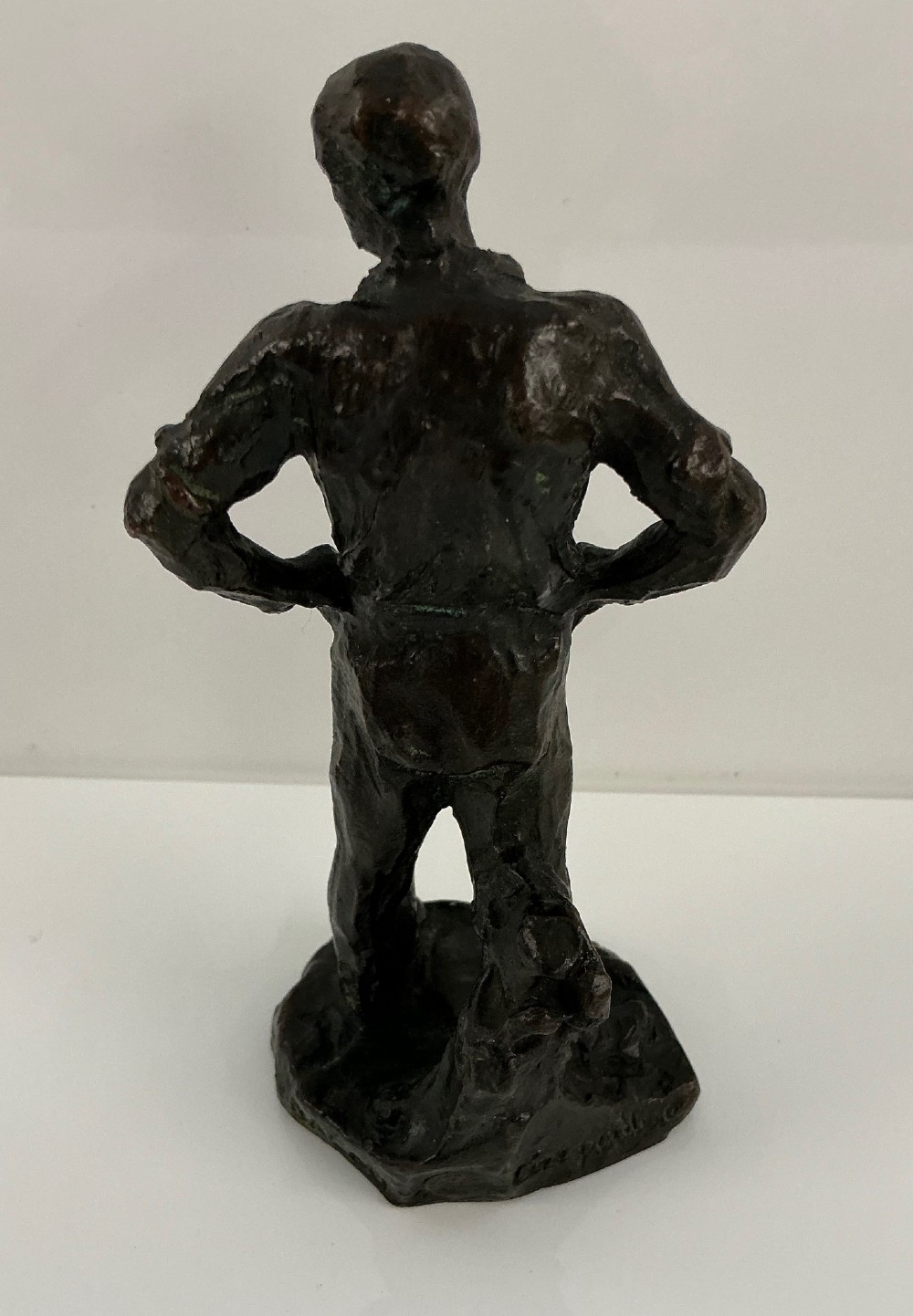 A dark patinated bronze sculpture depicting a standing male worker with his hands resting on his hips. View II