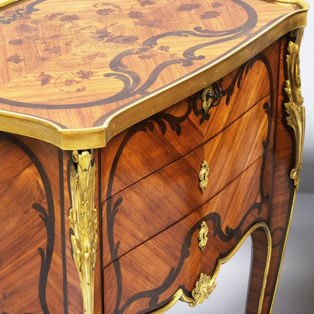 A pair of French Louis XV style inlaid timber commodes featuring serpentine fronts, marquetry veneering, cabriole legs, and chased gilt-bronze mounts. View IV