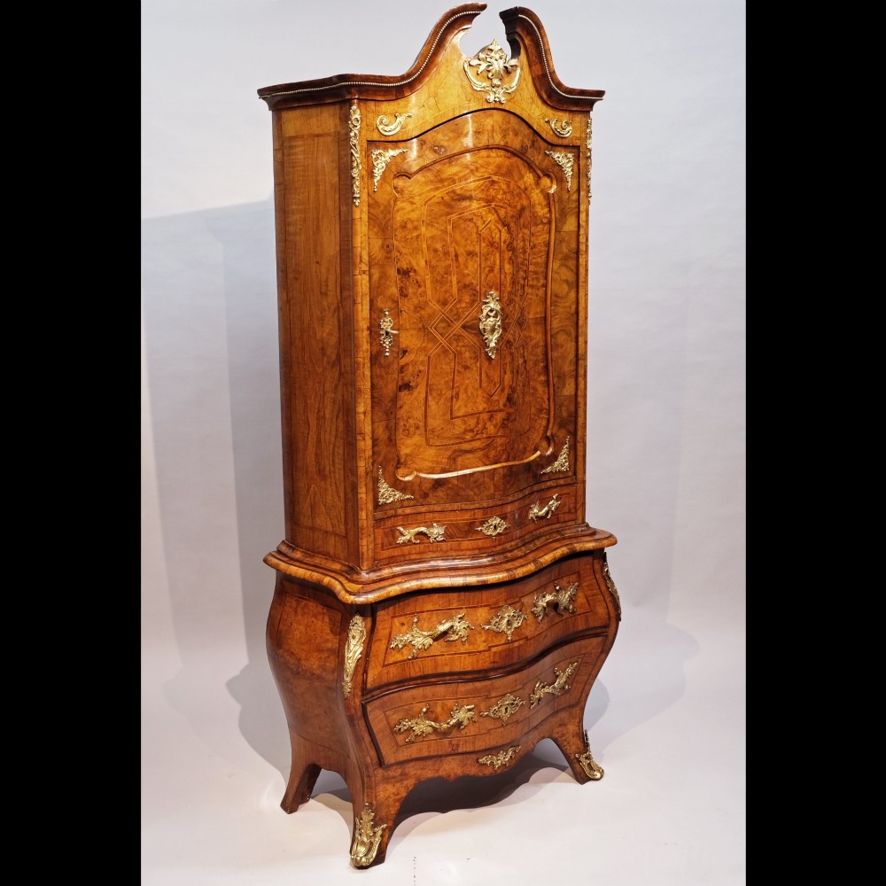 An antique 19th-century burr walnut cabinet on a bombe chest of drawers featuring ornate gilt-bronze mounts and an arched pediment top.
