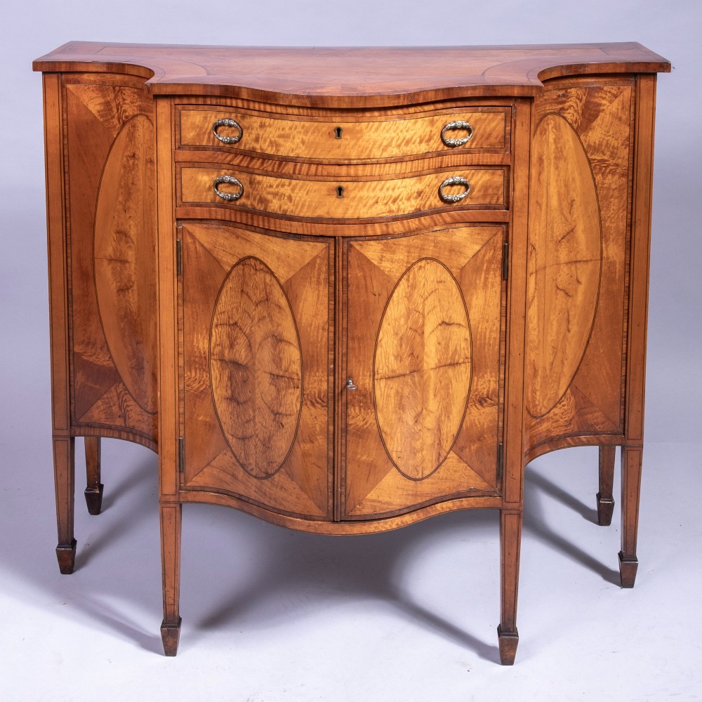A Sheraton Revival satinwood side cabinet featuring bow-fronted drawers, inlaid oval door panels, and tapered square legs.