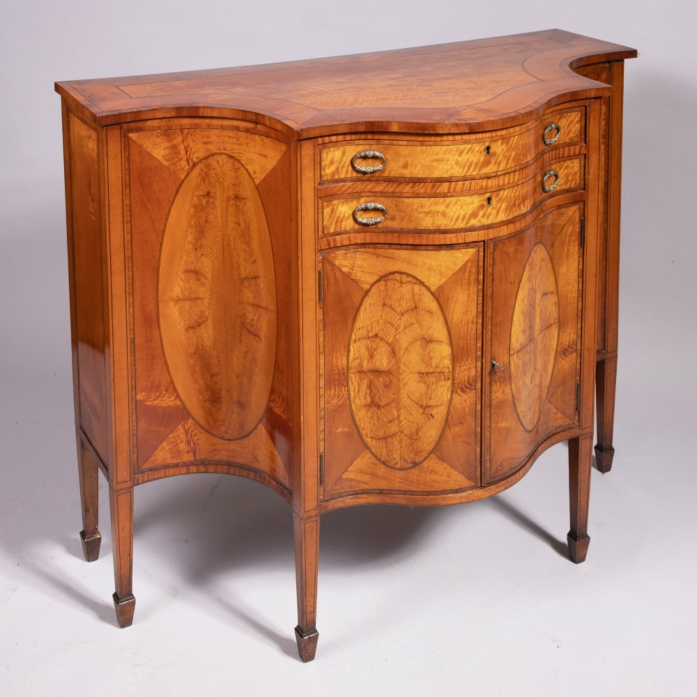 A Sheraton Revival satinwood side cabinet featuring bow-fronted drawers, inlaid oval door panels, and tapered square legs. View III