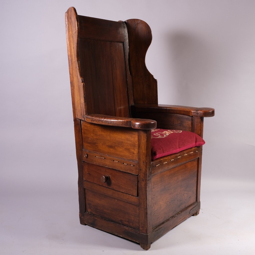 An eighteenth-century dark oak lambing chair with a high panelled back, side wings, box base with a single drawer, and a red upholstered seat cushion.