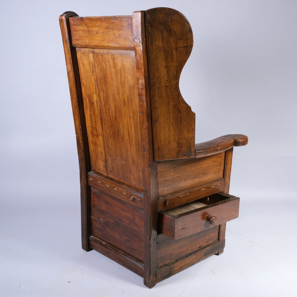 An eighteenth-century dark oak lambing chair with a high panelled back, side wings, box base with a single drawer, and a red upholstered seat cushion. View II