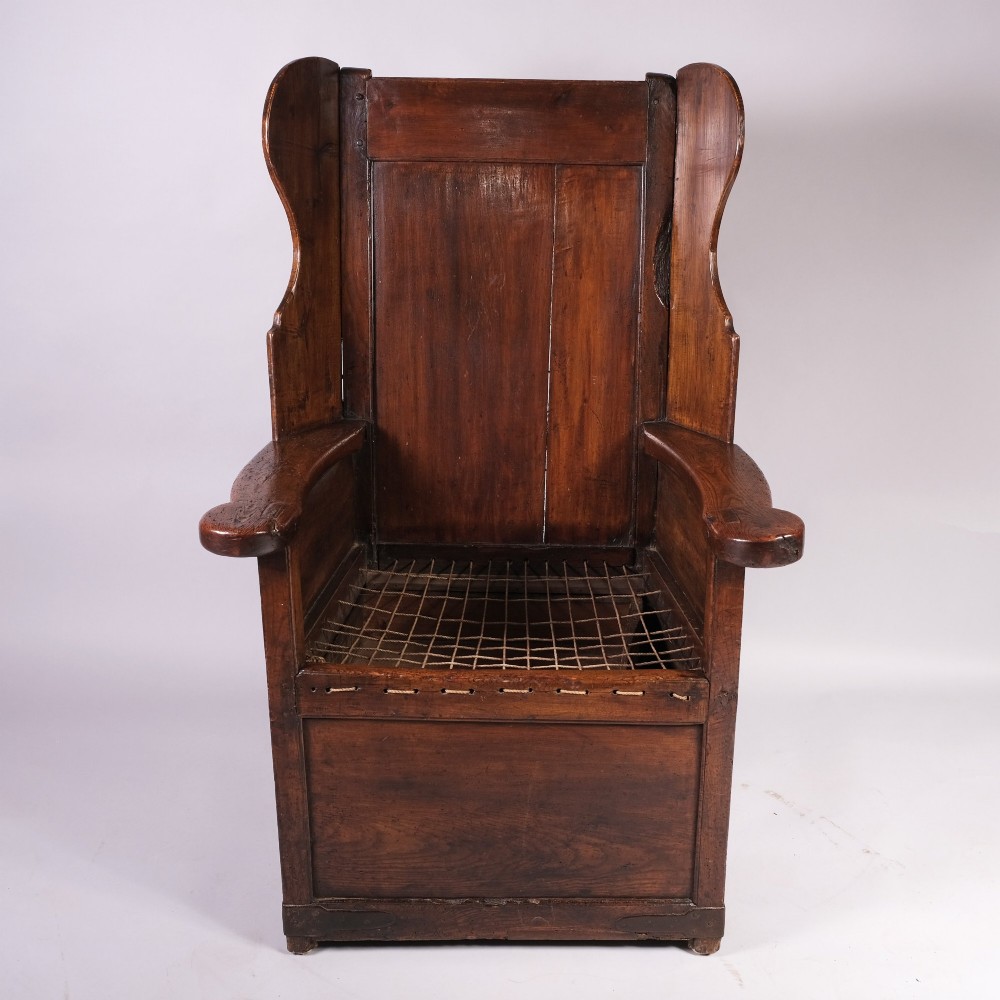 An eighteenth-century dark oak lambing chair with a high panelled back, side wings, box base with a single drawer, and a red upholstered seat cushion. View III