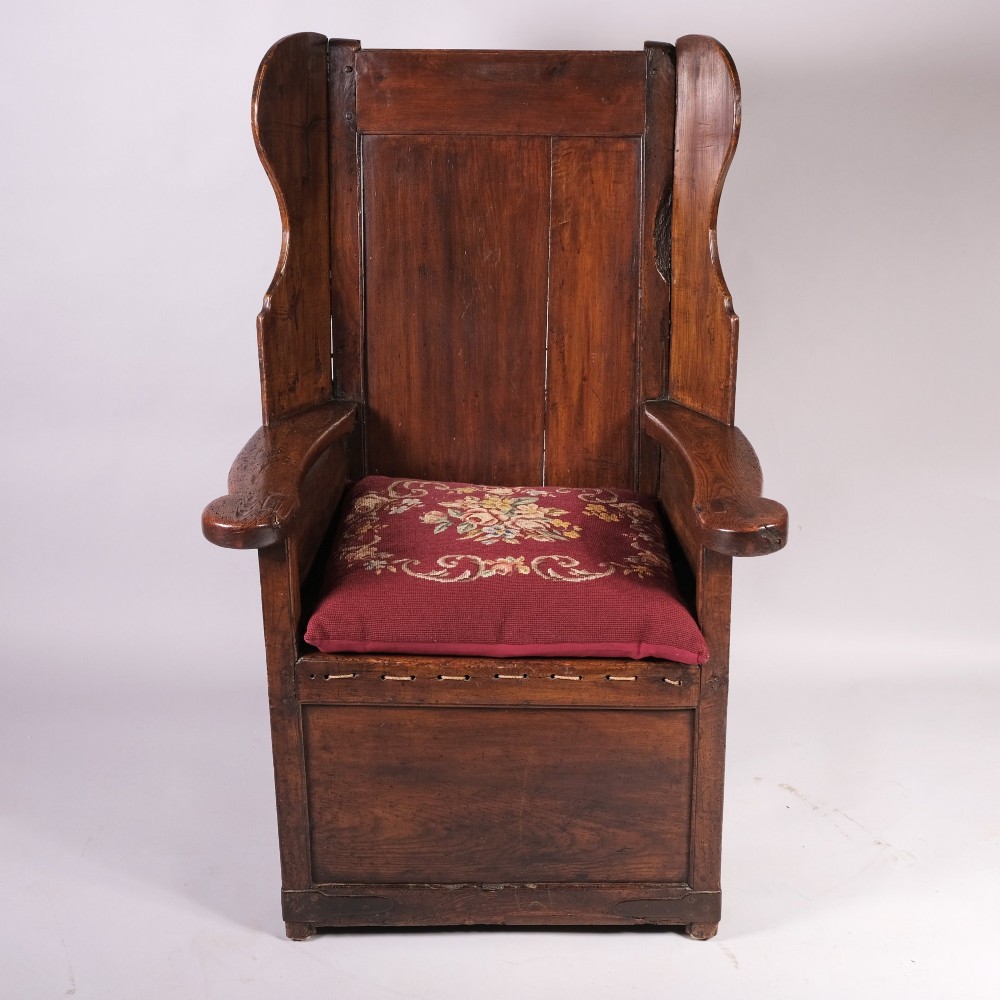 An eighteenth-century dark oak lambing chair with a high panelled back, side wings, box base with a single drawer, and a red upholstered seat cushion. View IV