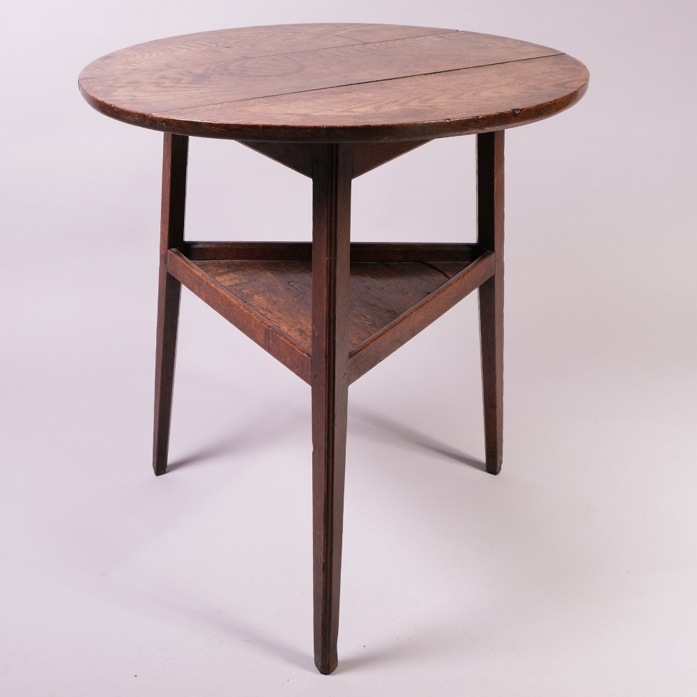 An 18th-century ash wood cricket table featuring a round top, three straight tapered legs, and a triangular lower shelf in a warm brown finish.