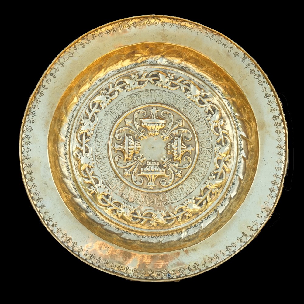 A circular 16th-century brass Nuremberg alms dish featuring central urn motifs surrounded by concentric bands of repousse scrollwork and Gothic script.