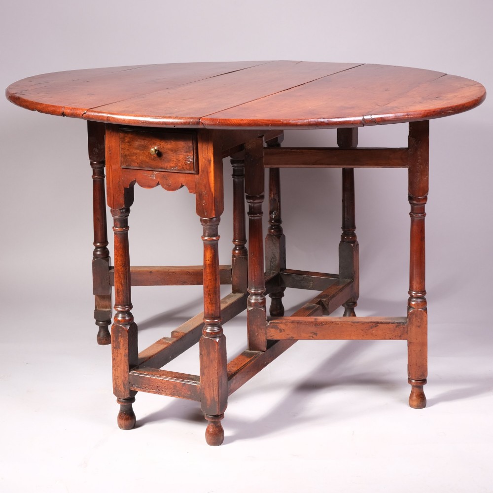 An 18th-century yew wood gateleg table featuring an oval drop-leaf top, turned legs, lower stretchers, and a single end drawer with a brass knob.