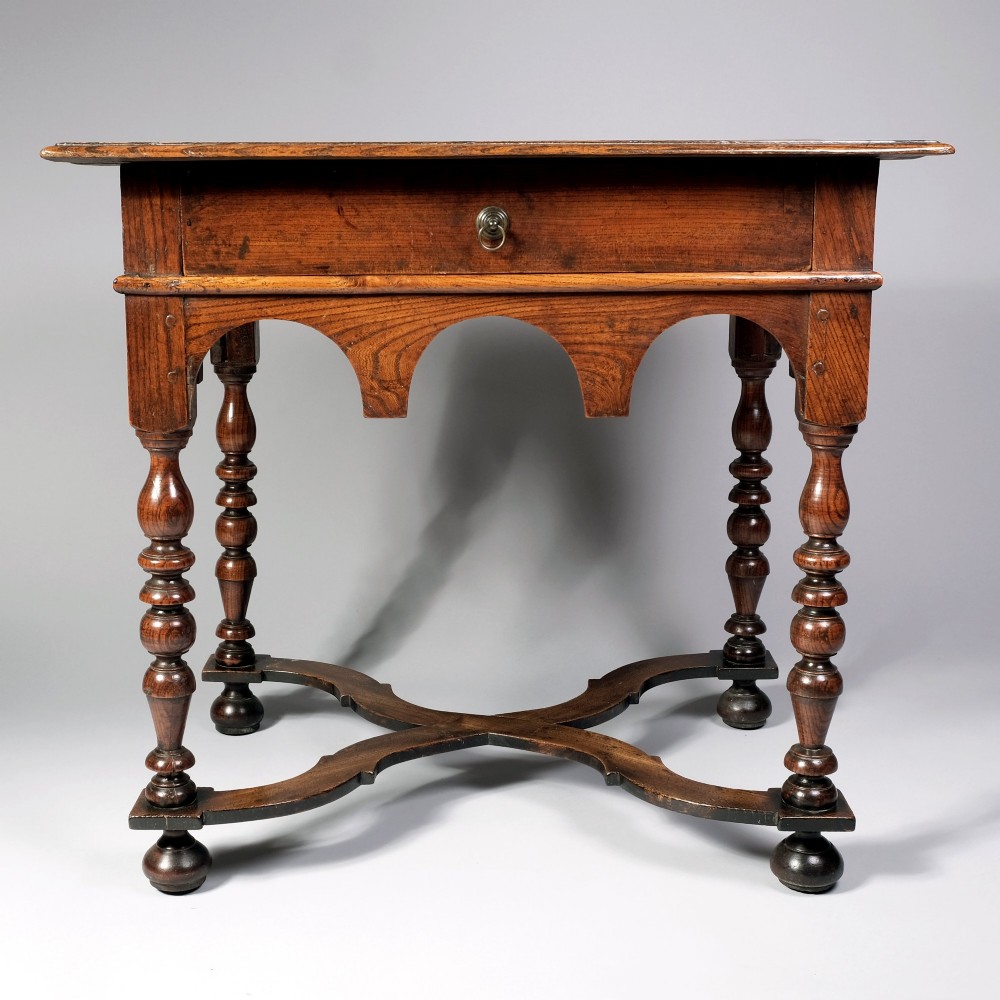 A late 17th century William and Mary period elm side table featuring turned legs, a single drawer, a shaped apron, and an X-frame stretcher.