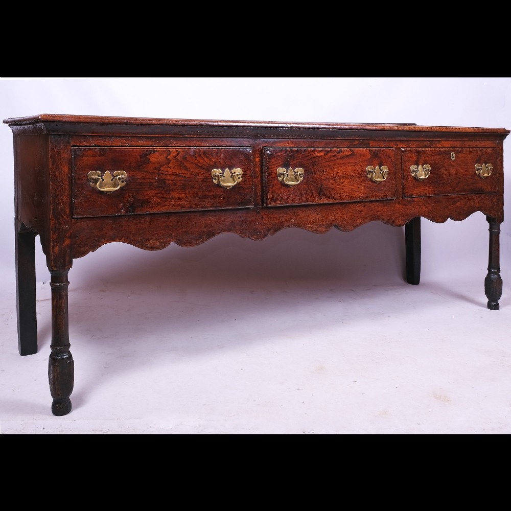 An early eighteenth-century Queen Anne dark oak dresser base featuring three inline drawers with brass handles, a scalloped apron, and turned front legs.