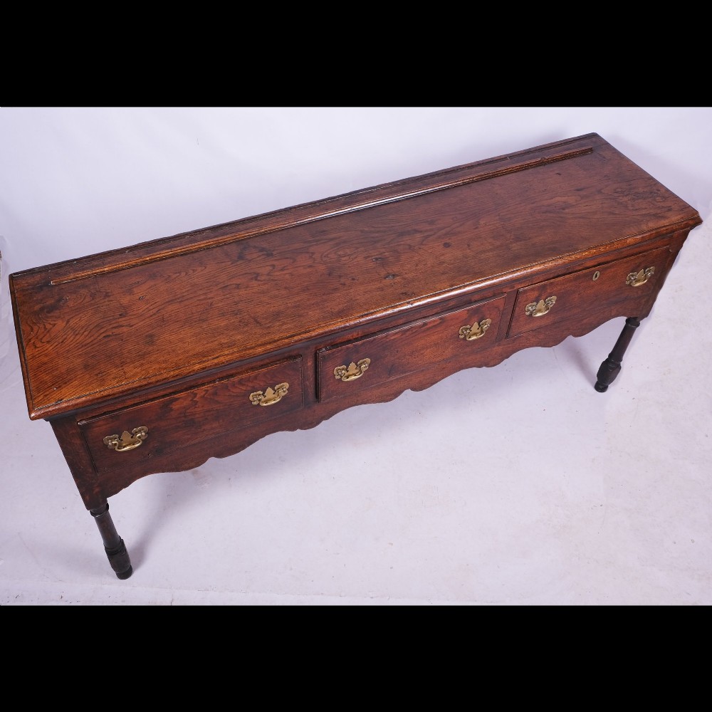 An early eighteenth-century Queen Anne dark oak dresser base featuring three inline drawers with brass handles, a scalloped apron, and turned front legs. View II