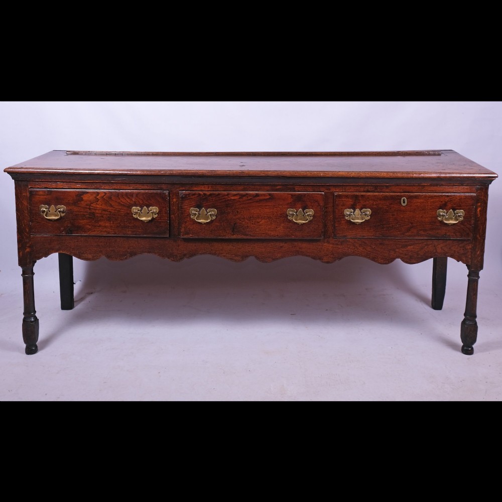 An early eighteenth-century Queen Anne dark oak dresser base featuring three inline drawers with brass handles, a scalloped apron, and turned front legs. View III