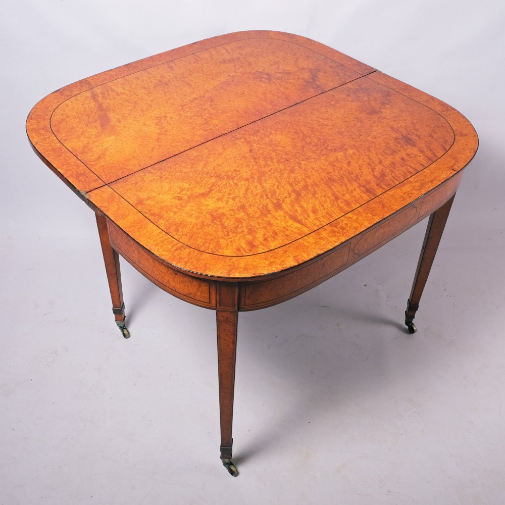 A late eighteenth-century Georgian amboyna fold-over tea table featuring string inlay, a rectangular apron with a central cartouche panel, and square tapered legs on brass castors. View II