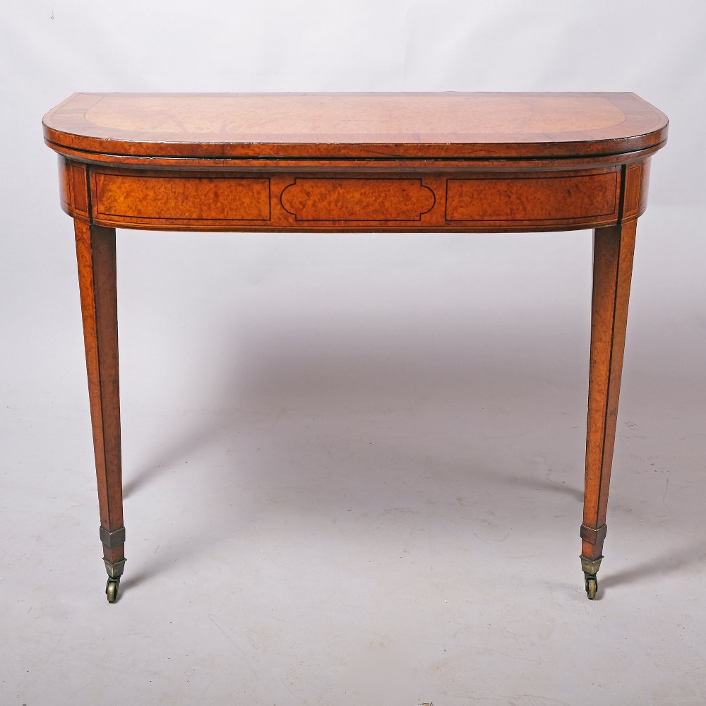 A late eighteenth-century Georgian amboyna fold-over tea table featuring string inlay, a rectangular apron with a central cartouche panel, and square tapered legs on brass castors. View IV