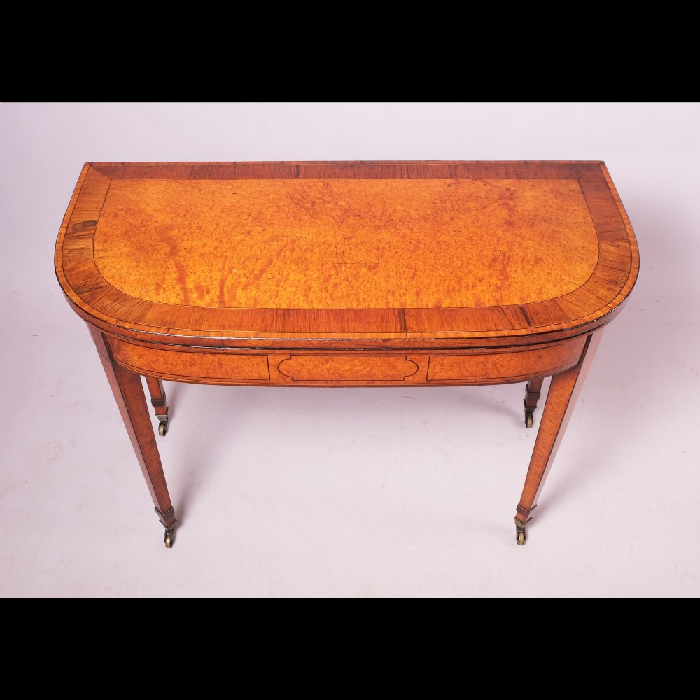 A late eighteenth-century Georgian amboyna fold-over tea table featuring string inlay, a rectangular apron with a central cartouche panel, and square tapered legs on brass castors. View V