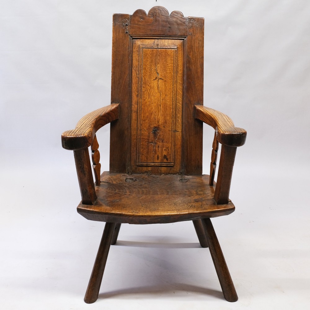 An antique mid-18th century dark oak panel-backed Windsor armchair featuring curved armrests, a shaped cresting rail, and splayed turned legs.