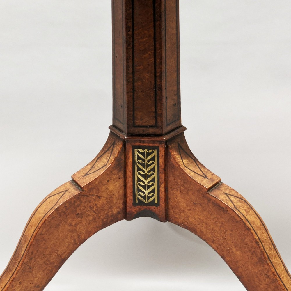 A Regency period oval tilt-top table crafted from amboyna wood with an ebony inlaid border, supported by a central pedestal and tripod legs with brass lion paw castors. View IV