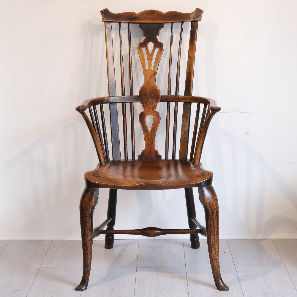 An 18th-century comb back Windsor armchair in warm brown timber, featuring a pierced central splat, spindle back, cabriole front legs, and a carved saddle seat.