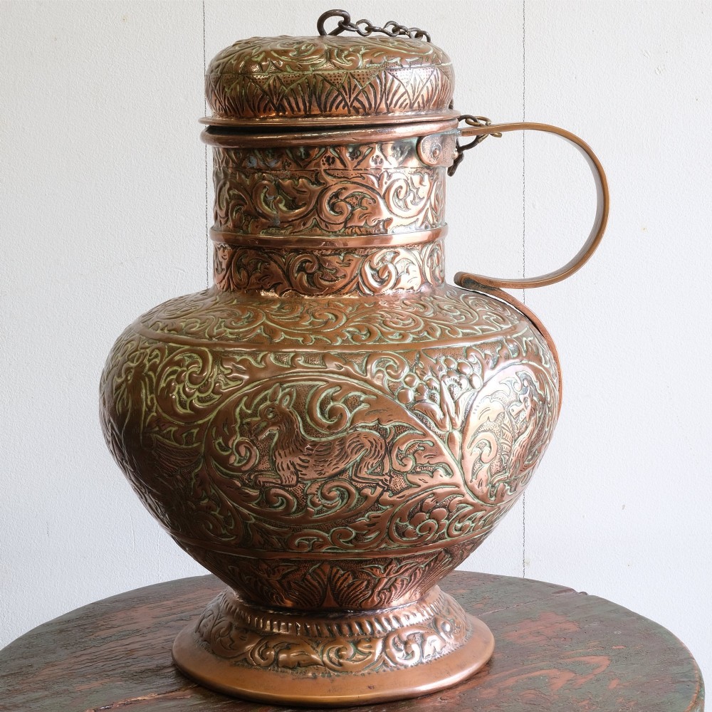A large 18th-century copper jug featuring hand-chased foliate scrollwork and animal motifs, complete with a domed chained lid and curved handle.