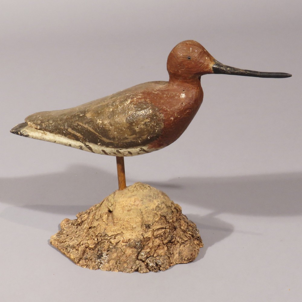 An antique hand-carved and painted wooden black-tailed godwit bird decoy with a long beak, mounted on a wooden rod set into a bark-covered base.