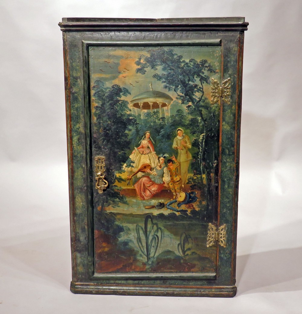 An early eighteenth-century wall-mounted corner cupboard crafted from dark green painted wood featuring a single door decorated with a hand-painted pastoral scene and pierced brass hinges.