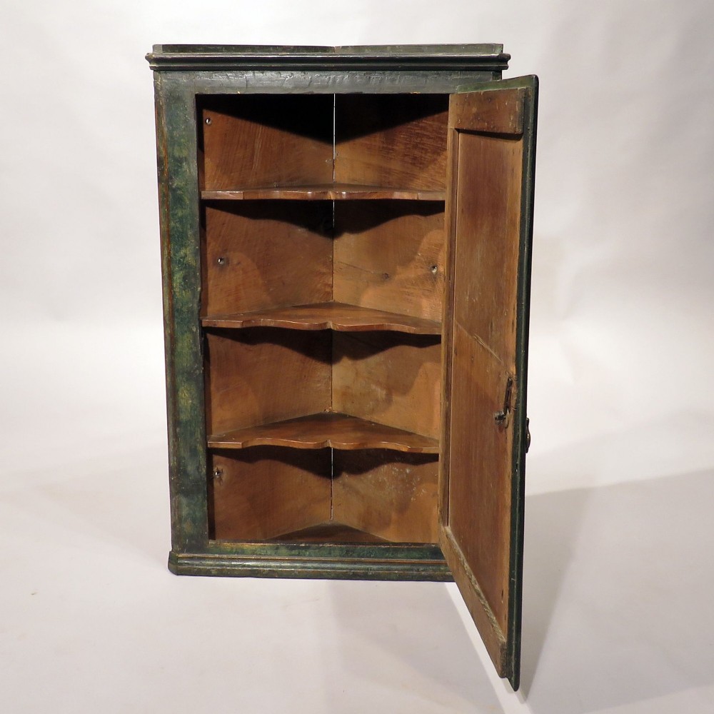 An early eighteenth-century wall-mounted corner cupboard crafted from dark green painted wood featuring a single door decorated with a hand-painted pastoral scene and pierced brass hinges. View II