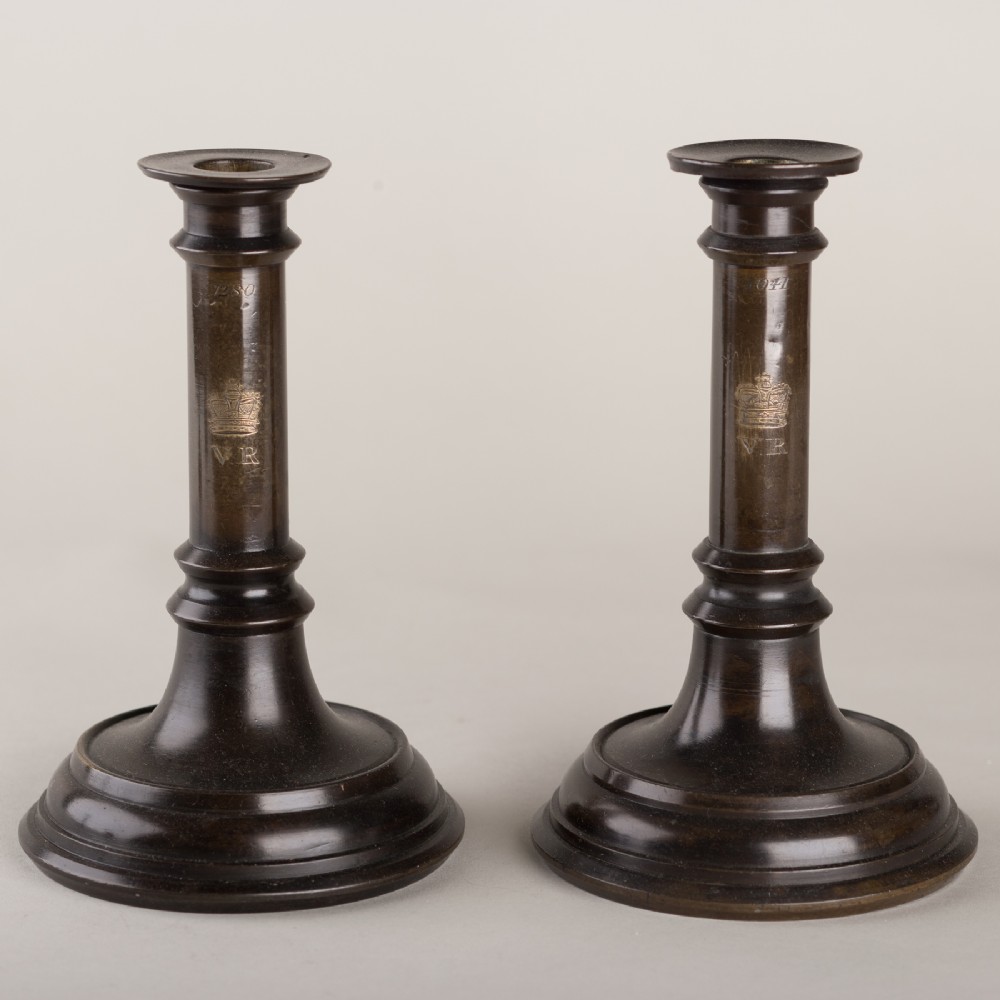 A pair of 19th-century dark patinated bronze candlesticks featuring turned circular bases and columns engraved with a royal crown and VR cypher.