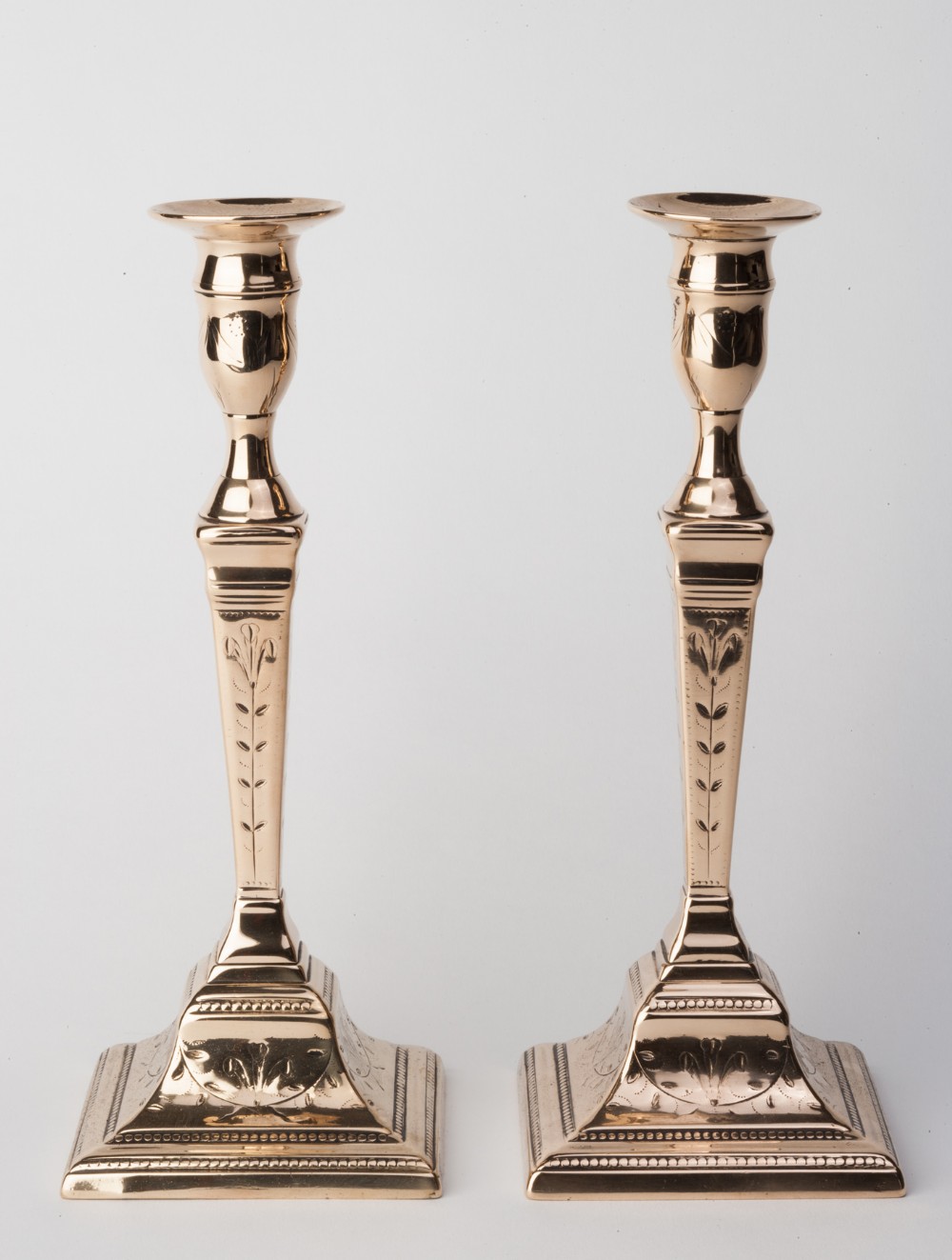 A pair of polished brass candlesticks featuring tapered square columns and detailed engraved stem motifs on stepped bases.