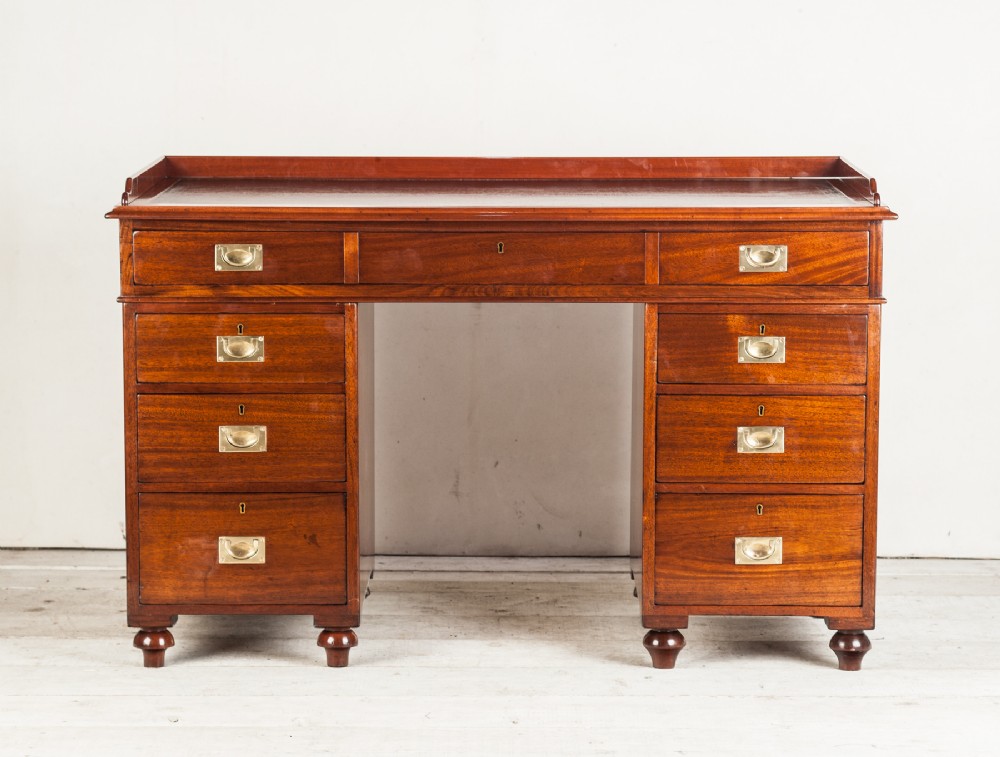 An antique mahogany military campaign pedestal desk featuring nine drawers with recessed brass handles, a gallery top, and turned feet.