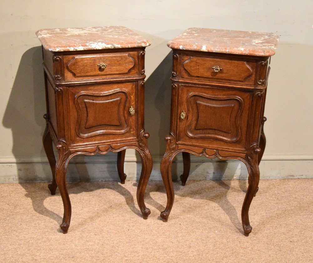 A pair of French carved oak bedside cabinets with variegated pink marble tops, single drawers, framed cupboard doors, and slender cabriole legs.