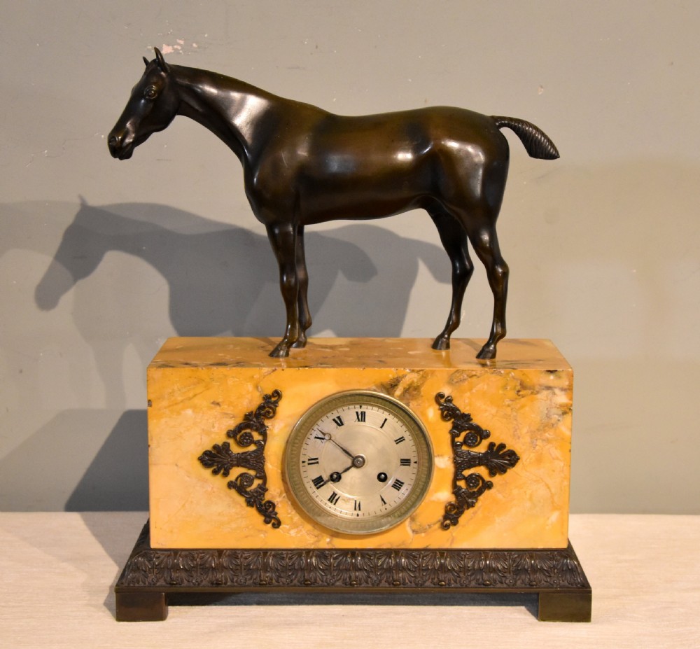 An antique mantel clock featuring a patinated bronze stallion standing atop a rectangular yellow Siena marble base with decorative scroll appliques and a circular dial with Roman numerals.