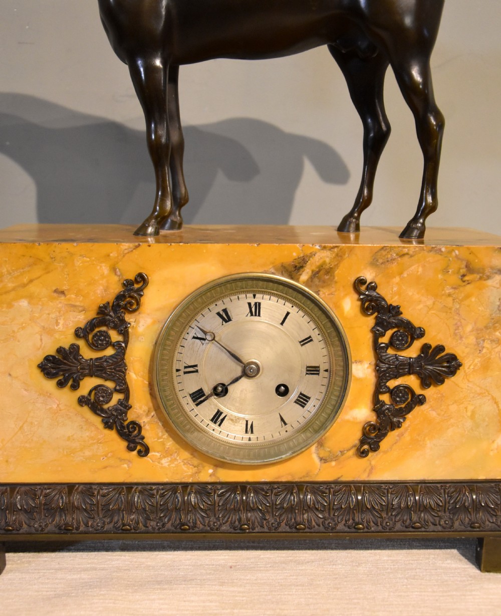 An antique mantel clock featuring a patinated bronze stallion standing atop a rectangular yellow Siena marble base with decorative scroll appliques and a circular dial with Roman numerals. View VI
