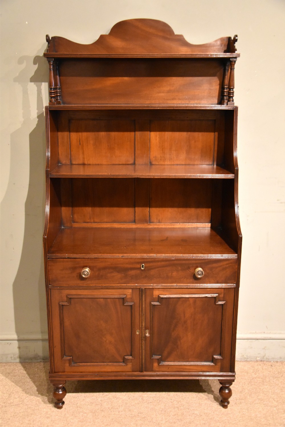 A Regency period mahogany waterfall bookcase featuring graduated open shelves, a single drawer, two lower cabinet doors, and turned feet.