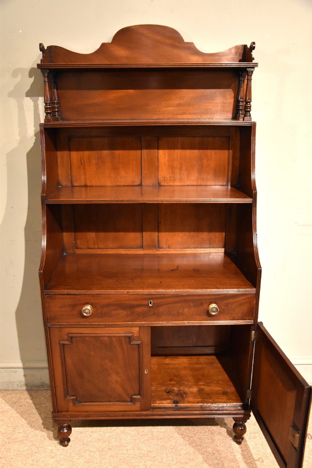 A Regency period mahogany waterfall bookcase featuring graduated open shelves, a single drawer, two lower cabinet doors, and turned feet. View II