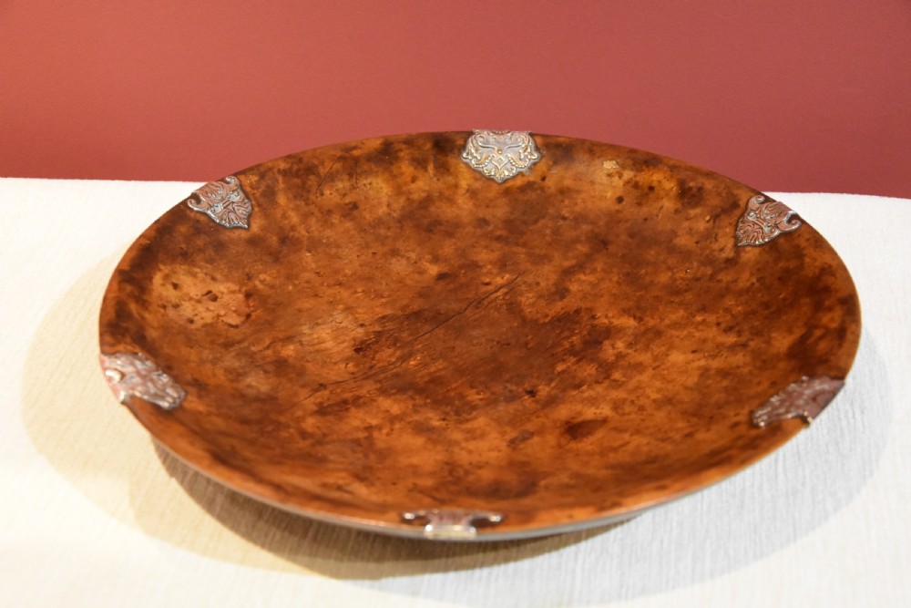 A circular shallow bowl carved from figured brown burr wood, featuring six chased silver mounts positioned around the rim. View IV