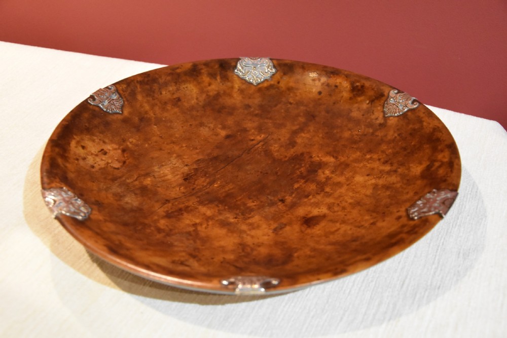 A circular shallow bowl carved from figured brown burr wood, featuring six chased silver mounts positioned around the rim. View V
