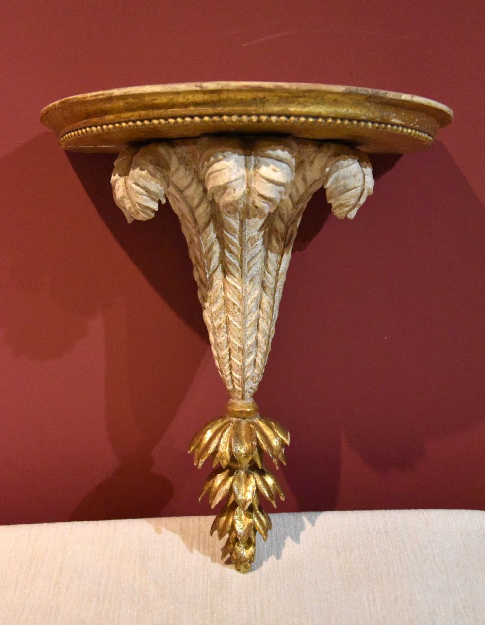 An 18th-century carved wall bracket featuring a semicircular gilded top shelf with beaded molding above a tapered white body with leaf carving and a drop leaf finial.